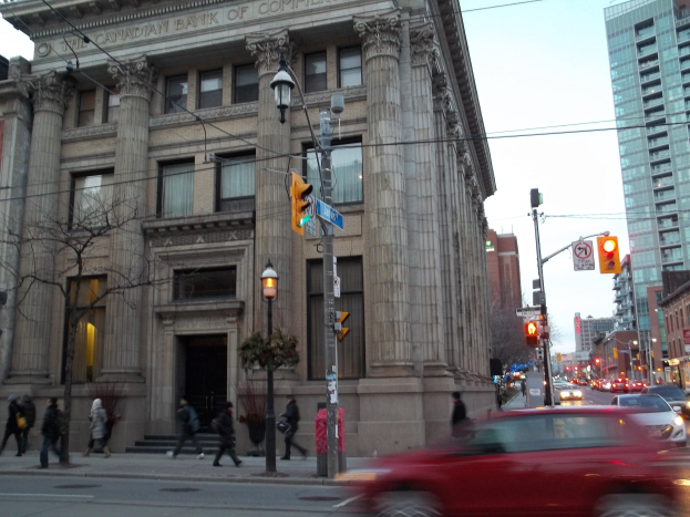 Eine belebte Stadtstraße mit Fahrzeugen, Fußgängern, Gebäuden und Bäumen, mit einem großen Gebäude im Vordergrund und einem roten Auto auf der Straße.