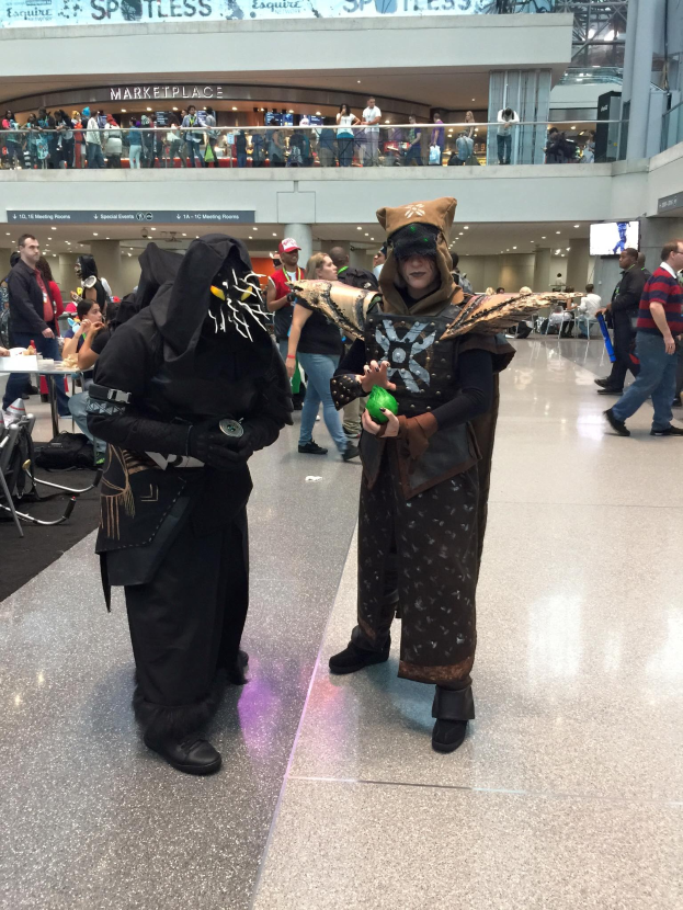 Zwei Personen in Kostümen stehen zusammen in einem belebten Flughafen, umgeben von sitzenden und stehenden Individuen, mit Geländern, Texttafeln, Lampen und anderen Gegenständen im Hintergrund.