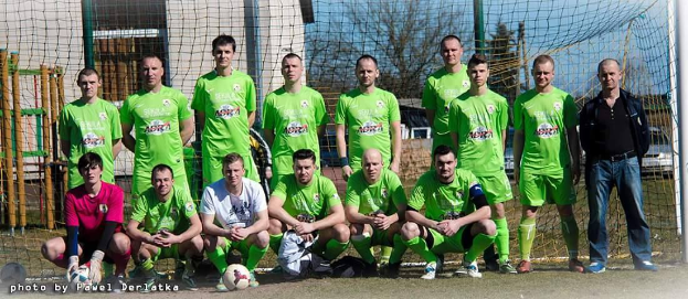 Eine Gruppe von Männern in grünen T-Shirts steht und hockt auf einem Fußballfeld mit einem Ball davor, einem Torpfosten rechts daneben und einer Hintergrundlandschaft aus Bäumen, Gebäuden, Pfählen, Fahrzeugen und Himmel.