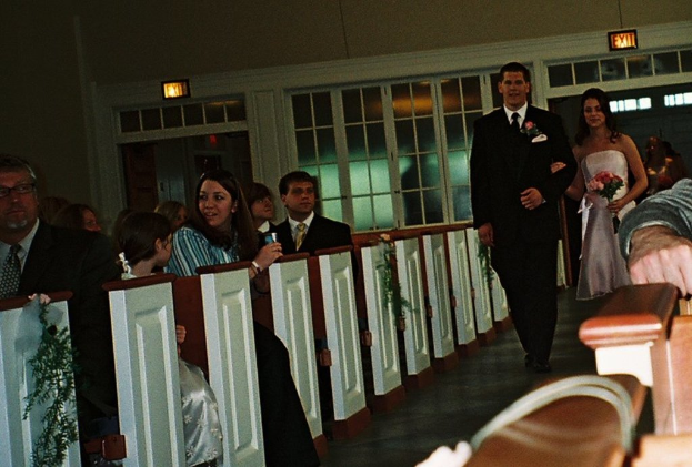 Eine Gruppe von Menschen sitzt auf einer Bank, während ein Brautpaar den Weg entlang auf sie zukommt, die Braut hält einen Blumenstrauß, mit Lampen und einem Fenster im Hintergrund, was auf eine Kirche hindeutet.