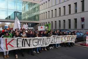Eine Gruppe von Menschen steht auf einer Straße und hält ein Banner mit der Aufschrift "Kein Grind Lo Feuer" hoch, mit Gebäuden, Bäumen, Polen, Schildern, Fahrzeugen und einem klaren blauen Himmel im Hintergrund.