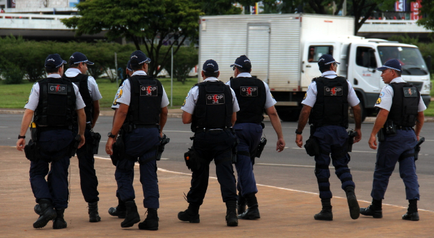 Eine Gruppe von Uniformierten, darunter Mützen, Hemden, Schutzwesten, Uhren, Hosen und Schuhe, die auf einer Straße mit einem geparkten Lkw auf der rechten Seite neben einem Zaun gehen. Im Hintergrund sind Gebäude, Bäume, Pflanzen, Pfosten und Banner zu sehen, mit einer Brücke in der oberen rechten Ecke.