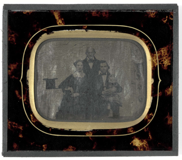 Schwarzes-Weiß-Foto eines Mannes in Anzug und Krawatte und einer Frau in einem unbekannten Outfit, die vor einem alten Fernseher stehen, beide mit ernsten Gesichtern.