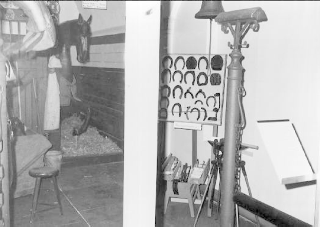 Schwarzes und weißes Foto eines Raums mit einem Pferd im Hintergrund, einer Person auf der linken Seite und verschiedenen Gegenständen wie einem Hocker und an der Wand angebrachten Rahmen, was auf einen Pferdestall hinweist.