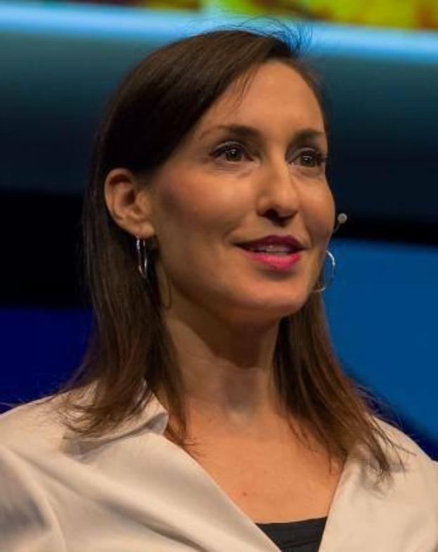 Frau in weißer Jacke und schwarzem Oberteil vor einem blauen und gelben Hintergrund sitzend, mit Ohrringen und Mikrofon während eines Interviews.