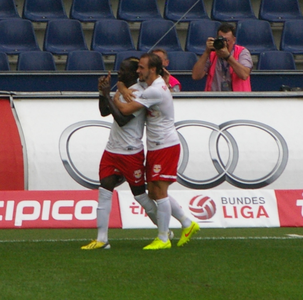 Zwei Fußballspieler feiern ein Tor auf einem Feld, umgeben von einer Gruppe von Menschen, mit einer Person mit einer Kamera, einem Banner mit Text, einigen Stühlen und einer Wand im Hintergrund.