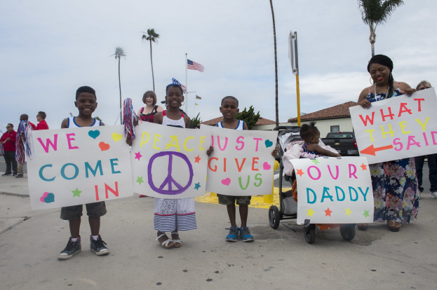 Eine Gruppe von Kindern hält Schilder vor einem Gebäude, während eine Frau einen Kinderwagen schiebt, die auf einer Straße umgeben von Bäumen, Pfählen, einer Flagge, einem Haus, Pflanzen und einem bewölkten Himmel stehen; auf einem Schild steht "Wir kommen in Frieden, gebt uns unseren Daddy."