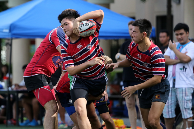 Eine Gruppe von Menschen, die draußen ein Spiel spielen, mit einem Jungen in der Mitte, der einen Ball hält, und Zuschauern im Hintergrund neben einem blauen Zelt.