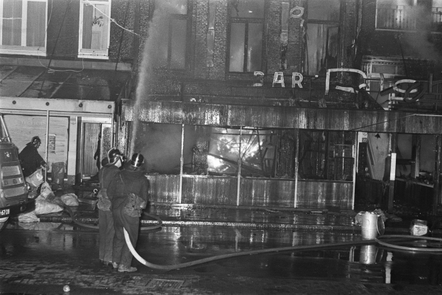 Feuerwehrleute bei der Brandbekämpfung an einer Straßenkreuzung, mit Menschen mit Rohren, einem geparkten Fahrzeug, verstreuten Gegenständen, einem Gebäude mit Fenstern, einem Schild mit Text und Kabeln unter einem bewölkten Himmel.