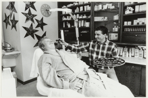 Ein Mann lächelt in einem Zahnarztstuhl, während ein anderer Mann neben ihm steht und eine Platte hält, mit einem Waschbecken, Sternen, einer Uhr und Regalen mit Flaschen im Hintergrund.