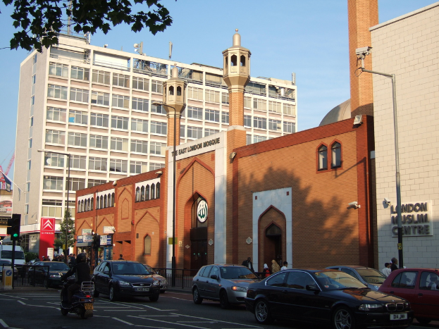 Eine Moschee mit Fenstern und Glastüren, die eine Wandtafel auf der Wand, Straßenlaternen, eine Ampel an einem Mast, Fahrzeuge auf der Straße, Gruppen von Menschen, die stehen, einen Baum und eine Überwachungskamera an einem Mast zeigt.