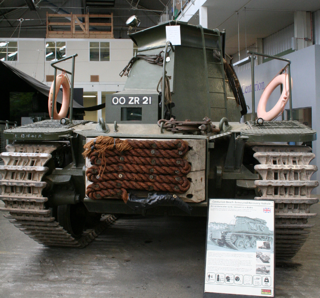 Ein Panzerfahrzeug ist auf dem Boden positioniert mit einer Tafel und mehreren Bildern in der Nähe, vor einem Hintergrund mit einer Wand, Säulen, Lampen, einer Hütte und Stangen.
