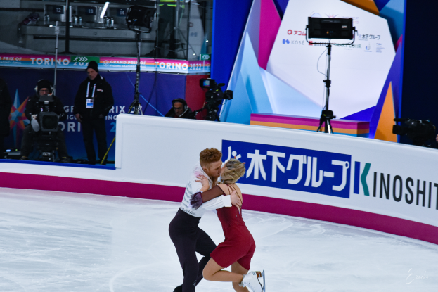 Ein Paar umarmt sich beim Eislaufen auf einer Eisbahn, im Hintergrund stehen einige Menschen, einige halten Kameras, und eine Tafel mit Text.