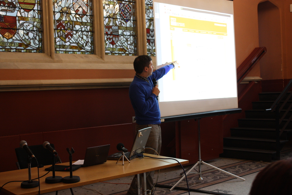 Ein Tisch mit Laptops, Kabeln und Mikrofonen in der Mitte, eine weiße Tafel mit einem stehenden Mann rechts, ein leerer schwarzer Stuhl hinter dem Tisch und im Hintergrund Treppen, Geländer, eine Wand mit Buntglas.