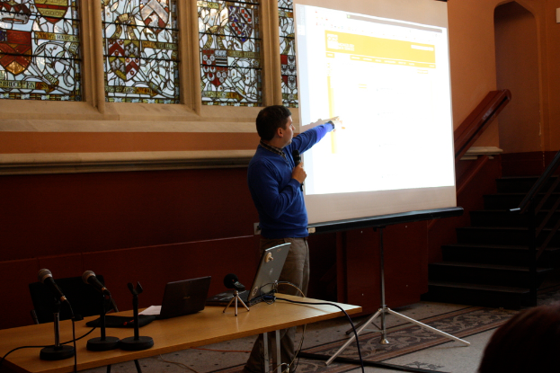 Ein Tisch mit Laptops, Kabeln und Mikrofonen in der Mitte, eine weiße Tafel mit einem stehenden Mann rechts, ein leerer schwarzer Stuhl hinter dem Tisch und im Hintergrund Treppen, Geländer, eine Wand mit Buntglas.