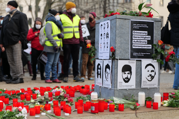 Eine Gruppe Menschen steht ernsthaft vor einem mit Kerzen, Blumen und Plakaten geschmückten Denkmal, umgeben von Bäumen und Gebäuden mit Fenstern im Hintergrund, und gedenkt einer Tragödie in Berlin, Deutschland.