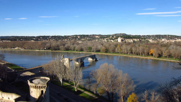 Eine Brücke überspannt einen Gewässserbereich, mit Fahrzeugen auf der Straße, umgeben von Bäumen, alter Architektur und Gebäuden, unter einem bewölkten Himmel.