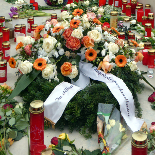 Ein Mahnmal in Berlin, Deutschland, mit einem Strauß Blumen und Kerzen, die auf dem Boden vor einer Wand aufgestellt sind und von Blättern und anderen Gegenständen umgeben sind.
