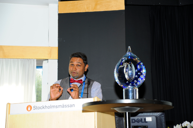 Ein Mann steht neben einem Rednerpult und spricht mit einer Wand im Hintergrund.