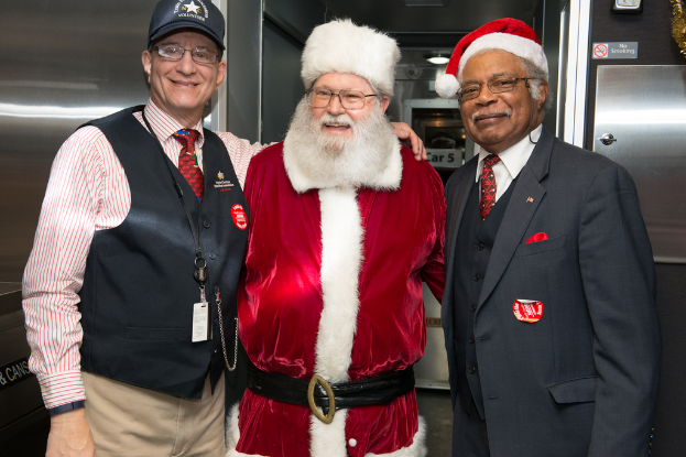 Eine Gruppe von Menschen in Brillen, Mützen, Krawatten, Ausweisen und Abzeichen, darunter eine Person in einem Weihnachtskostüm, vor einer Stahlwand mit Lichtern, Schildern und anderen Gegenständen im Hintergrund.
