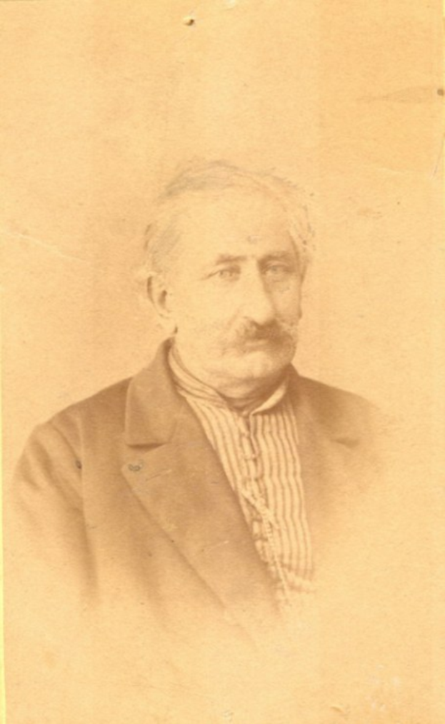 Ein Schwarz-Weiß-Foto eines älteren Mannes mit Schnurrbart, der einen Mantel trägt und ein starkes Kinn, eine markante Nase, dunkle Augen und einen nachdenklichen Ausdruck mit leicht geöffneten Lippen hat.