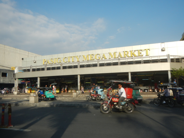 Eine belebte Straßenszene am Pasig City Mega Market mit Fahrzeugen und Fußgängern, einem Gebäude mit Text im Hintergrund und einem bewölkten Himmel.