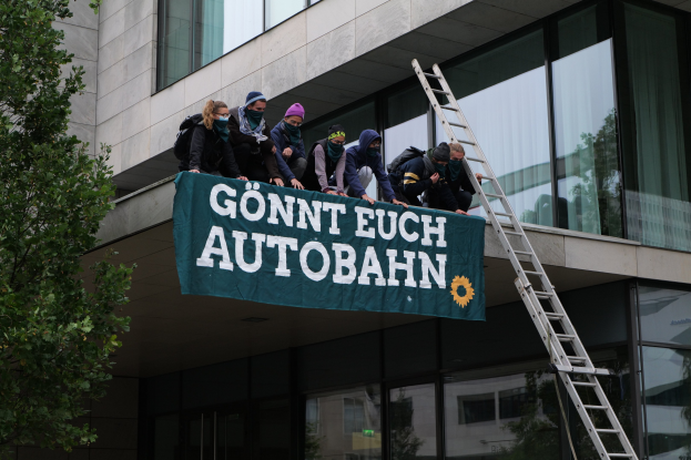 Gruppe von Menschen auf einem Dach, die eine Fahne halten, mit einem Baum links und Glasfenstern im Hintergrund, wahrscheinlich protestieren sie für die deutsche Regierung.