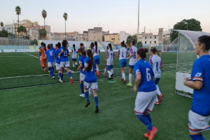 Gruppe von Frauen beim Fußballspielen auf einem Outdoor-Feld mit Gebäuden, Bäumen und einem Torpfosten im Hintergrund.