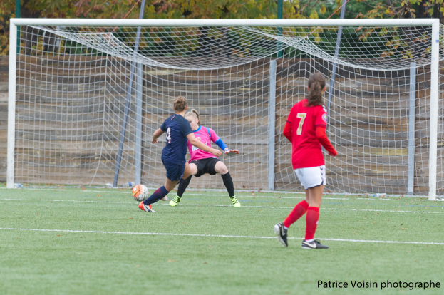 Eine Gruppe von Frauen beim Fußballspielen auf einem grünen Rasenfeld umgeben von Bäumen, mit einem Tor im Hintergrund und Text am unteren Bildrand.