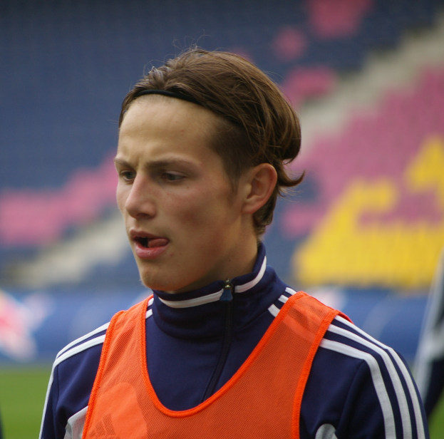 Ein junger Mann in einer orangen Weste steht auf einem Fußballfeld, die Arme ausgestreckt, mit einem entschlossenen Gesichtsausdruck, während der Hintergrund unscharf ist.
