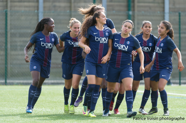 Gruppe junger Frauen, die auf einem Rasenfußball spielen, mit Maschendrahtzaun und Wand im Hintergrund, Text unten rechts: "Paris Saint-Germain Women's Soccer".