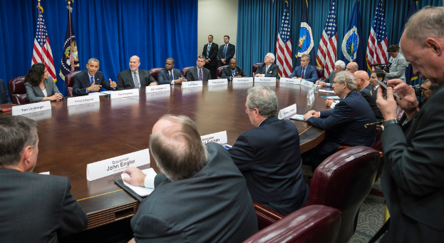 Eine Gruppe von Menschen, die an einem Tisch sitzen, mit einigen, die in der Ecke stehen, Fahnen vor einem blauen Vorhang.