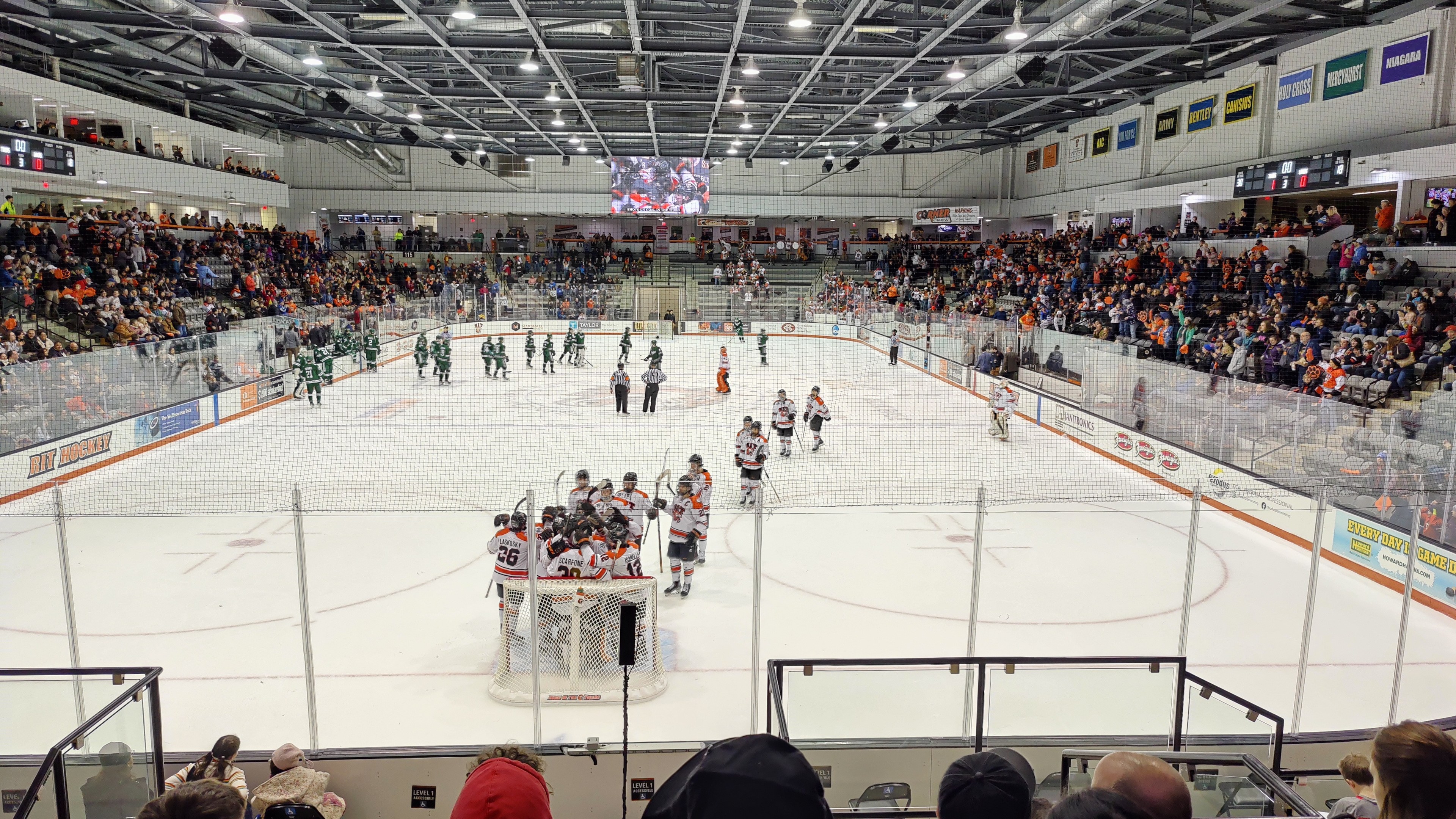 Ein Hockey-Spiel in einer Eishalle mit Spielern, Netz, Zuschauern in Tribünen, Hallenbeleuchtung, Bildschirm und Banner an der Decke.