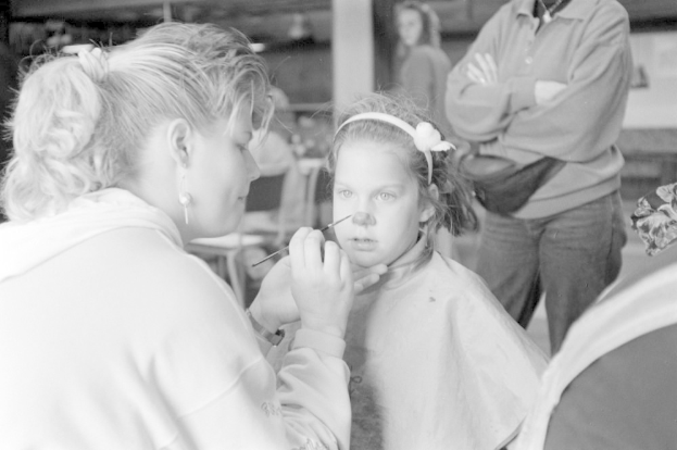 Ein junges Mädchen lässt sich von einer Frau in einem Salon Make-up auflegen, während zwei andere Personen im Hintergrund stehen, alles in Schwarz-Weiß dargestellt.