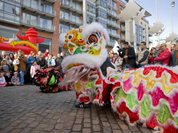 Ein lebendiges chinesisches Neujahrsfest in Amsterdam mit einem Drachen tanzen im Vordergrund und einer Menge Menschen, einige halten Kameras, umgeben von Gebäuden, Laternenmästen und einem klaren blauen Himmel.