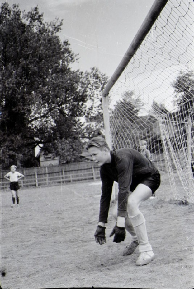 Schwarz-weiß-Foto eines jungen Jungen, der auf einem Feld mit einem Zaun, Bäumen und einem klaren Himmel Fußball spielt, mit einer Person in Schuhen im Vordergrund und anderen Kindern, die auf dem Boden spielen.