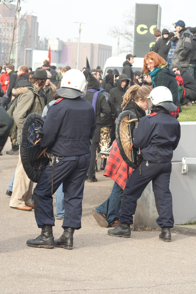 Eine Gruppe von Menschen auf einer Straße, mit zwei Personen in der Mitte, die wie Polizeibeamte aussehen, Gebäuden im Hintergrund und Boden unten.