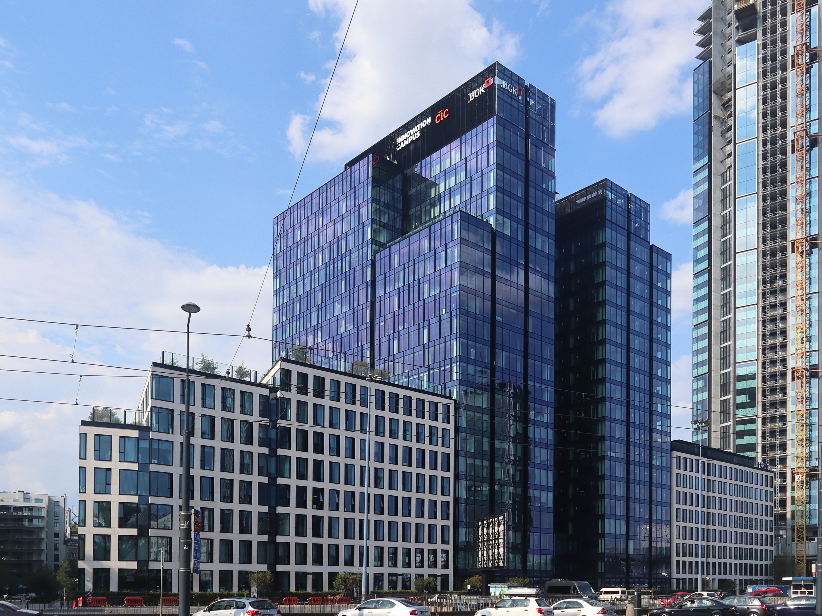 Zentrale der Deutschen Bank in Frankfurt, Deutschland, ein großes Gebäude mit vielen Fenstern, umgeben von städtischen Elementen einschließlich Straßenlaternen, Fahrzeugen, Grünflächen und einem bewölkten Himmel.