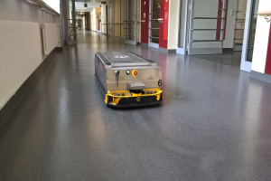 Ein Roboter, wahrscheinlich ein Reinigungsgerät, steht auf dem Boden eines Gebäudes und ist von Wänden, Türen, Lampen und anderen Gegenständen umgeben.