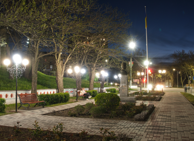 Ein Park bei Nacht mit einem beleuchteten Weg, der von Laternen, Bänken, Pflanzen, Gras, Bäumen und einem Fahnenmast mit einer Fahne gesäumt ist, mit Gebäuden und einem Sternenhimmel im Hintergrund.