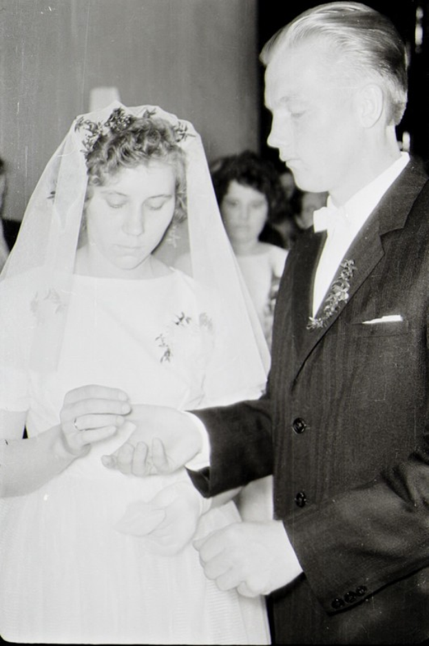 Ein Schwarz-Weiß-Foto einer Braut in einem weißen Kleid und Schleier und eines Bräutigams in einem Anzug, die sich ihre Eheringe austauschen, mit ein paar Leuten im Hintergrund.