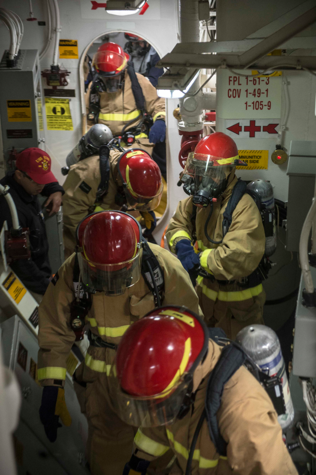 Eine Gruppe von Feuerwehrleuten in Schutzausrüstung, einschließlich Helmen, Handschuhen und Sauerstoffzylindern, arbeitet an einem Boot mit Rohren und anderen Gegenständen im Hintergrund und Plakaten mit Text an der Wand.