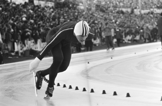 Ein Schwarz-Weiß-Foto eines Eisschnellläufers in Aktion auf dem Eis, der einen Helm und Eislaufschuhe trägt, mit ein paar unscharfen Menschen im Hintergrund.