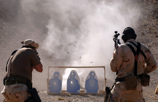 Zwei Männer in Tarnuniformen schießen mit Gewehren auf ein Ziel, mit Rauch in der Luft und einem hölzernen Objekt auf der rechten Seite.