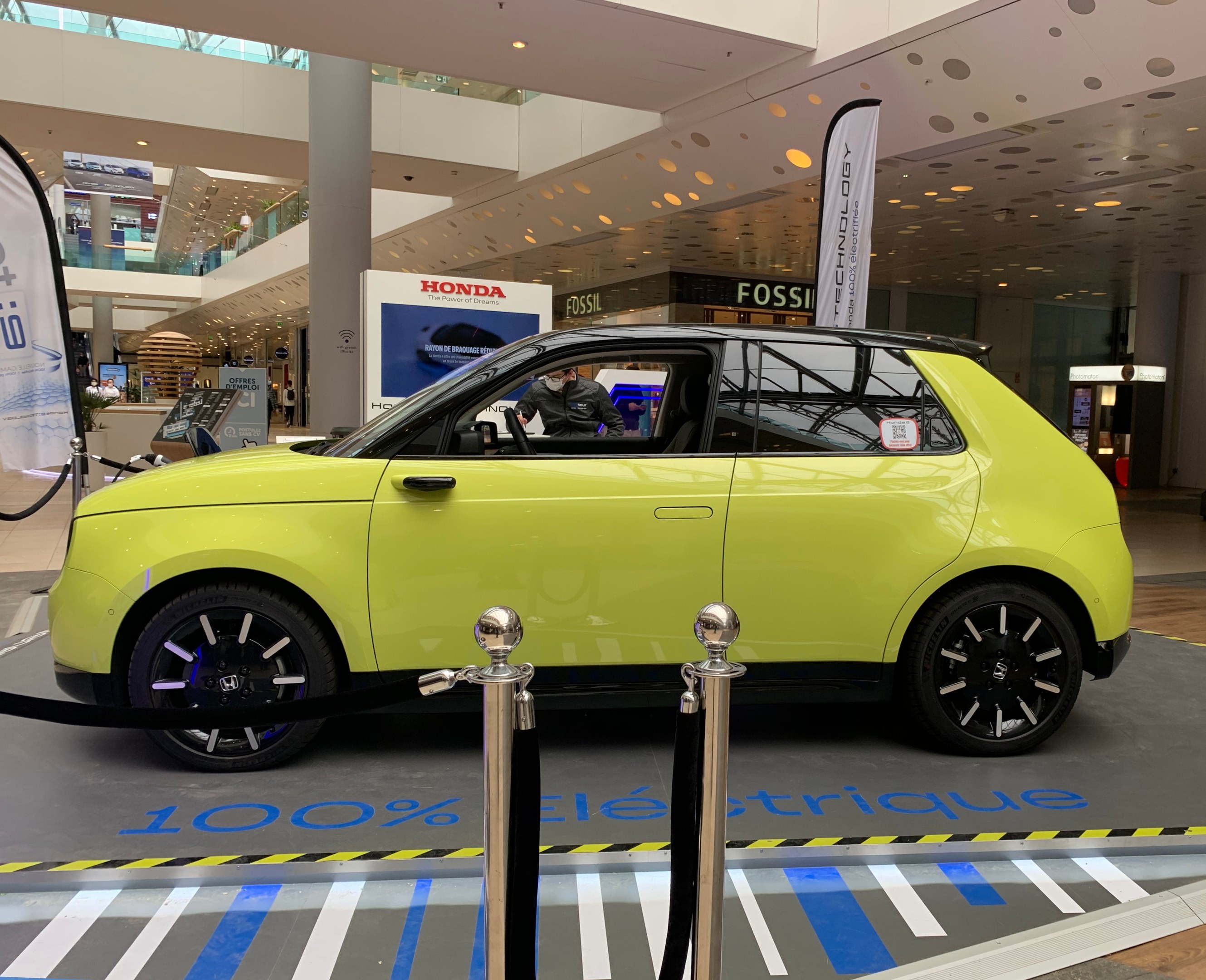 Ein gelbes Wasserstoff-elektrisches Auto, das vor einem Gebäude geparkt ist, umgeben von Barrieren und Bannern, mit Menschen in der Nähe und einer Pflanze in einem Topf.