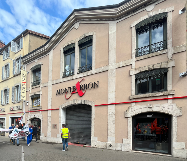 Eine Gruppe von Menschen mit Fahnen geht eine Straße entlang vor einem Gebäude mit einer Tafel, die 'Montbron' lautet, einem Straßenschild und einer Fahne mit Bildern und Text, unter einem bewölkten Himmel.
