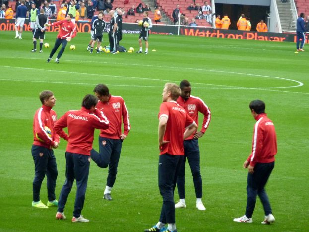 Eine Gruppe von Fußballspielern, die auf einem grünen Feld mit Fußballschuhen stehen und ein Publikum im Hintergrund auf Stühlen sitzt.