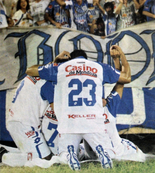 Gruppe von Fussballspielern, die auf dem Boden vor einer jubelnden Menge feiern, mit einem Banner mit Text im Hintergrund.