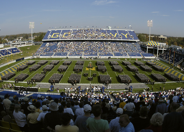 Eine große Menge schaut ein Fußballspiel in einem Rasenstadion unter einem klaren blauen Himmel, mit Laternenmasten, Bäumen und Gebäuden im Hintergrund.