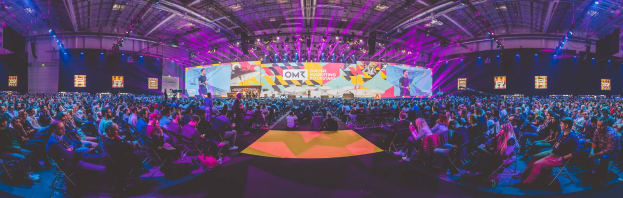 Eine große Menschenmenge sitzt auf Stühlen vor einer hell erleuchteten Bühne mit mehreren stehenden Personen, wahrscheinlich bei einem Online Marketing Rockstars Event, umgeben von Bildschirmen.